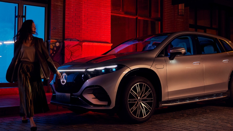 <p>Vista lateral delantera de Camioneta Eléctrica Mercedes-Benz EQS SUV color gris estacionada en la calle con una mujer al frente caminando.</p>