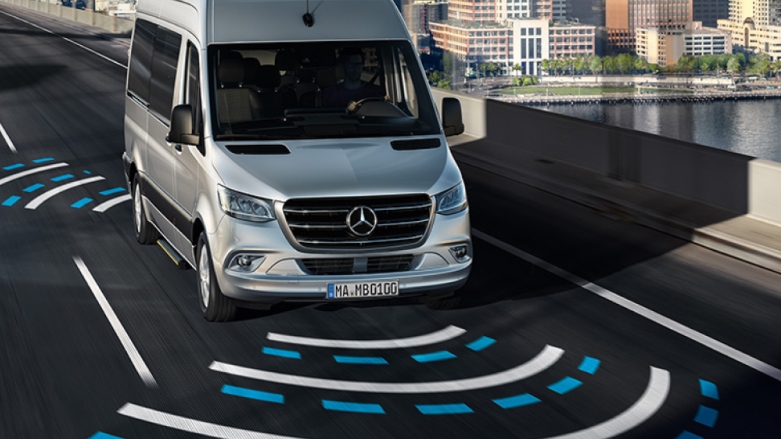 Van de lujo Mercedes-Benz Sprinter de pasajeros color gris siendo conducida por calles de la ciudad mostrando su tecnología de programa de estabilidad adaptativo. 
